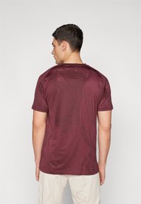 Camiseta deportiva color burdeos con un sutil patrón geométrico, mangas cortas, un cuello redondo y un tejido texturizado en la parte posterior.