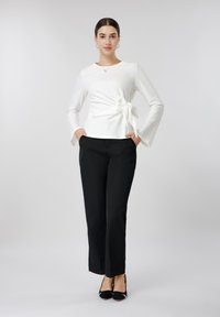 Γυναίκα στάζει με τα χέρια στις τσέπες, φοράει λευκό μπλουζάκι με μακριά μανίκια και δεσίματα στο πλάι, μαύρο παντελόνι ίσιας γραμμής και μαύρα τακούνια.
