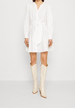Witte jurk met lange mouwen, een V-hals, knoopsluiting en een strikceintuur in de taille. Gecombineerd met glanzende beige kniehoge laarzen.