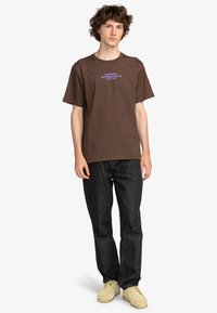 Brown cotton t-shirt with a round neckline and short sleeves featuring purple text "ELEMENT SKATEBOARD CO. WORLD WIDE." Paired with dark jeans.