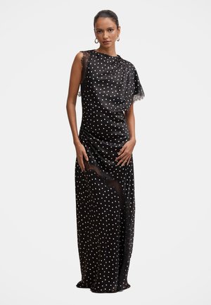 Mujer con un vestido negro sin mangas con lunares blancos y detalles de encaje, de pie con las manos cerca de las caderas sobre un fondo liso.