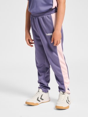Kind draagt paarse trainingsbroek met roze strepen aan de zijkant en witte sneakers met zwarte chevron-logo's, staand op een effen achtergrond.
