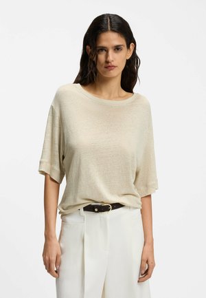 Frau mit dunklem, welligem Haar trägt ein locker sitzendes, beiges Kurzarmoberteil, das in weiße, hochtaillierte Hosen mit einem schwarzen Gürtel gesteckt ist.