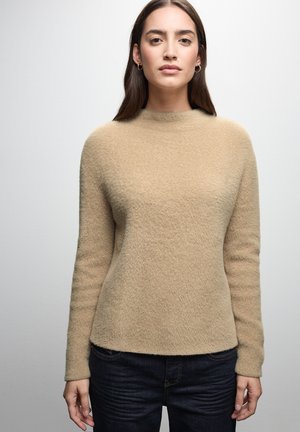 Maglione beige realizzato in un morbido tessuto testurizzato con un collo alto. Le maniche lunghe presentano un delicato motivo a righe. Abbinato a jeans scuri.