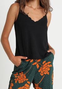 Donna che indossa un top nero a scollo a V con bordi scallop e pantaloni verdi con grande stampa floreale arancione e pois bianchi, con le mani nelle tasche.