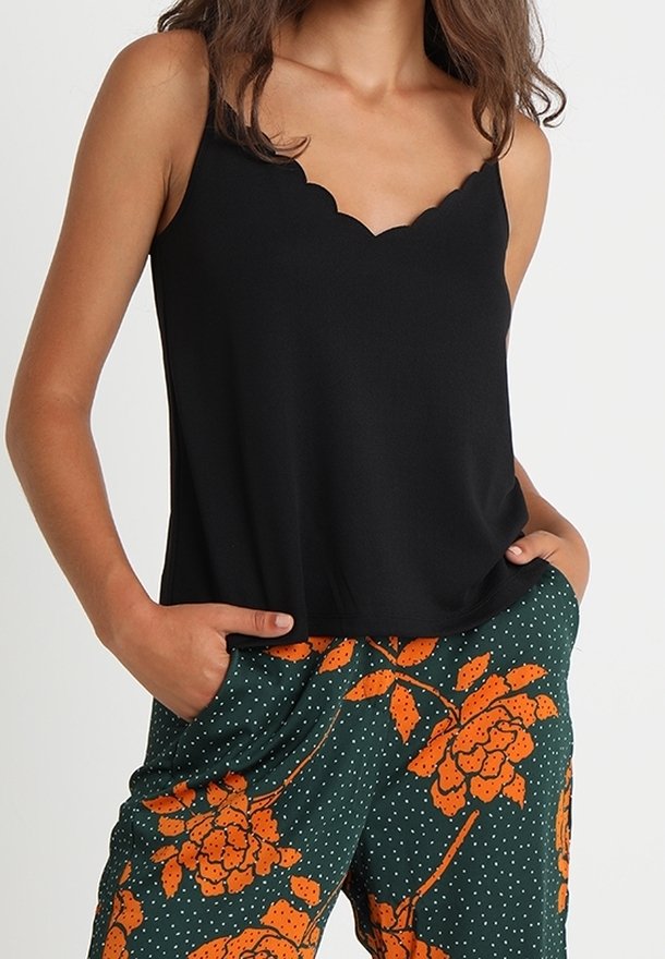 Donna che indossa un top nero a scollo a V con bordi scallop e pantaloni verdi con grande stampa floreale arancione e pois bianchi, con le mani nelle tasche.