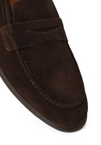 Mocasín de ante marrón oscuro con detalle de tira de moneda cosida y puntera redondeada, visible sobre un fondo blanco.