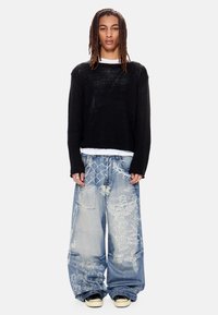 Άτομο με μακριά dreadlocks που φοράει μαύρο πουλόβερ, λευκή μπλούζα κάτω από το πουλόβερ, φαρδιά ανοιχτό μπλε τζιν με λευκά σχέδια αγγέλων και μαύρα αθλητικά παπούτσια.