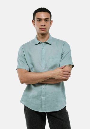 Chemise à manches courtes vert clair en tissu texturé, avec un devant boutonné et une seule poche poitrine, portée avec un jean foncé.