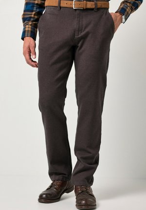 Mann trägt dunkelbraune Hose mit beigem Gürtel, blau-braunes kariertes Hemd, braune Lederschuhe, eine Hand in der Tasche, steht vor neutralem Hintergrund.