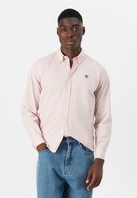 Light pink button-down shirt with long sleeves, made of soft fabric, featuring a small logo on the left chest. Paired with blue jeans.