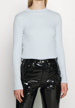 Persoon die een lichtblauw nauwsluitend shirt met lange mouwen draagt, ingestopt in een hooggetailleerde, glanzende zwarte vinylbroek met knopen aan de voorkant en zakken.