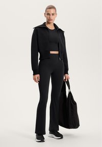 Fekete cropped zipzáras dzseki, hozzá illő testhez simuló felső és harangszárú leggings. Kiegészítők között szerepel egy nagy fekete túra táska és fekete edzőcipők.