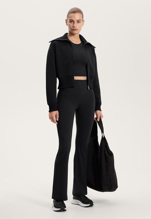 Sort cropped zip-up jakke, matchende tætsiddende top og flare leggings. Tilbehøret inkluderer en stor sort tote bag og sorte løbesko.