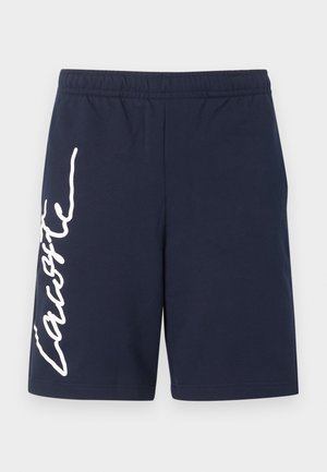Marineblå shorts med elastisk midje og hvit vertikal "Lacoste"-skrift på venstre ben, ensfarget design, midt på låret.