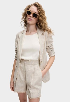 Beige linneblazer och shorts set med trasiga kanter, kombinerat med en vit topp och stora fyrkantiga solglasögon. Slät tygtextur.