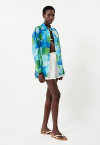Farbige, übergroße Button-Up-Bluse im blau-grünen Karomuster, kombiniert mit weißen Denim-Shorts und beigen Flip-Flops. Glattes Material.