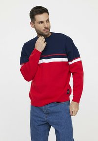 Pull en tricot rouge et marine avec des rayures horizontales blanches et marines. Doté d'un col rond et de poignets côtelés. Porté avec un jean en denim bleu.