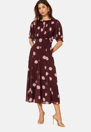 Naine kannab burgundia midi kleiti heledate roosade täppidega, lühikeste tülliliste varrukatega, liibuvat vööd ja beeže kõrge kontsaga sandale.