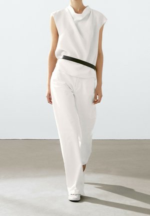 Person walking wearing white sleeveless top and wide-leg white pants with a black belt, against a plain light background.