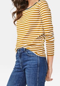 Gul- och vitrandig långärmad topp med svart halsringning, kombinerad med blå denimjeans. Tyget verkar mjukt och åtsittande.