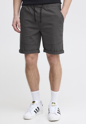 Homme portant un short gris foncé à ourlet roulé avec taille à cordon, chaussettes blanches montantes et baskets Adidas blanches à rayures noires, debout.
