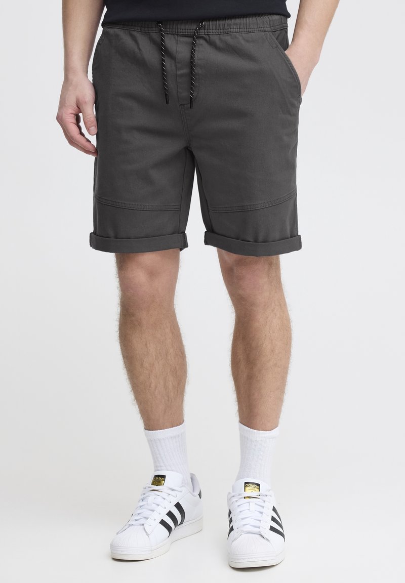 Homme portant un short gris foncé à ourlet roulé avec taille à cordon, chaussettes blanches montantes et baskets Adidas blanches à rayures noires, debout.