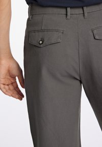 Un pantalon gris présente une coupe slim, des passants de ceinture, et deux poches arrière avec des fermetures à bouton. Le tissu semble lisse et léger.