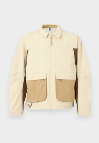 Beige a hnědá bunda se zipem vpředu, velkými předními kapsami a límcem. Vyrobena z lehkého materiálu, s logem na hrudi.