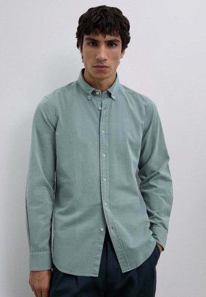 Camisa verde abotonada hecha de un material suave que presenta un cuello y mangas largas. Se observa una textura sutil y un pequeño detalle de logotipo.