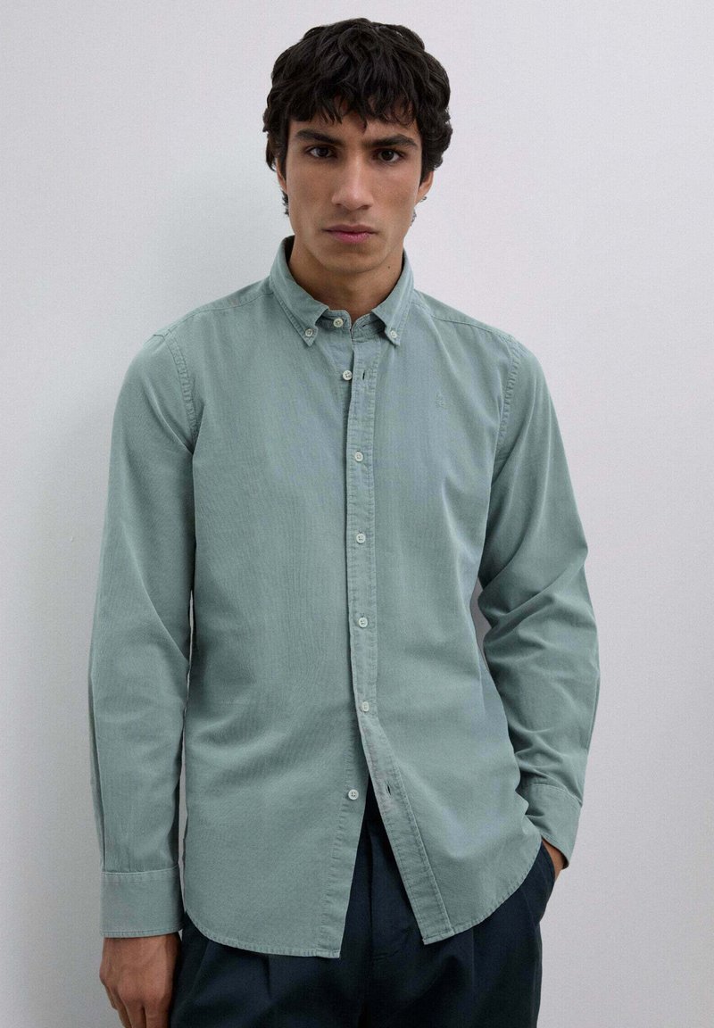 Grøn knappet skjorte lavet af et blødt materiale har en krave og lange ærmer. En subtil tekstur og lille logo detalje er synlig.