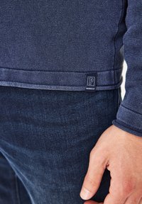 Marineblauer Pullover aus strukturiertem Stoff mit gerippten Bündchen, versehen mit einem kleinen blauen Etikett mit dem "Paddock's"-Logo nahe dem Saum, getragen über dunkler Jeans.