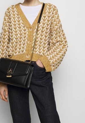 Žlutý a bílý pletený kardigan se vzorem a velkými knoflíky, nošený přes bílou košili, doplněný tmavými džínami a černou kabelkou přes rameno.