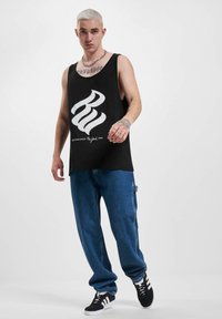 Čierny bezrukávový top s veľkým bielym grafickým dizajnom. Nosí sa s voľnými modrými džínsy a čiernymi teniskami s bielymi pruhmi.