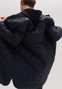 Prošívaná námořnická bunda s kapucí, která má diamantový vzor, hladkou texturu a zapínání na knoflíky a zip. Otevřená, aby ukázala vnitřní podšívku.