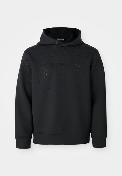 Črna kapuca z teksturirano površino in srebrnimi okrasnimi žebljički. Spredaj prikazuje logo "Emporio Armani" v kontrastnem slogu. Mehka tkanina.