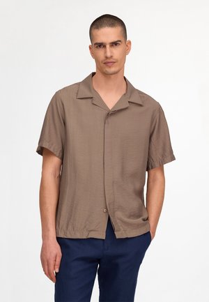 Mann mit kurzen Haaren, der ein locker sitzendes braunes kurzärmeliges Hemd mit Knöpfen und dunkelblaue Hose vor einem einfarbigen Hintergrund trägt.