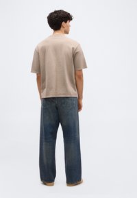 Νεαρός άνδρας με σγουρά μαλλιά, φορώντας μπεζ κοντομάνικη μπλούζα και μπλε τζιν παντελόνι φαρδιάς γραμμής, στέκεται με την πλάτη προς το μέρος μας σε απλό φόντο.