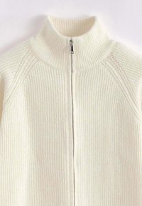 Un pull zippé en tricot côtelé crème avec un col haut, présentant une fermeture éclair centrale et un design en tissu texturé, idéal pour le superposition.
