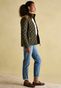 Jakna u maslinasto zelenoj boji s kvadratnim uzorkom i svijetlosmešnim naglascima na ramenima, visokim kragnom, vertikalnim patentnim zatvaračima, u kombinaciji s svijetloplavim farmerkama i smešnim oksfordicama.