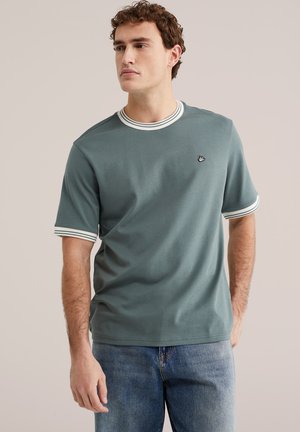 Jeune homme portant une chemise verte à manches courtes avec un col et des poignets rayés blancs, associée à un jean bleu, regardant vers la gauche.