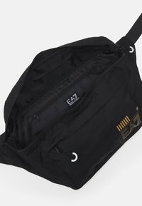 Borsa a spalla nera in tessuto con un ampio scomparto principale, tasca con zip, dettagli in metallo bianco e accenti logo dorati sul davanti.