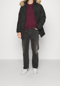 Schwarze Parka-Jacke mit pelzgefütterter Kapuze, burgunderfarbener Pullover, dunkle Jeans und weiße Turnschuhe. Die Jacke verfügt über eine Knopfleiste und seitliche Taschen.