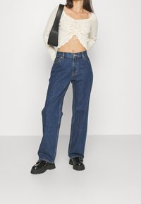 Lehký pletený crop top s vlnitým lemem a šněrováním, kombinovaný s volnými tmavě modrými džíny a robustními černými botami.