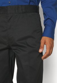 Svarta byxor med en slät textur och en platt framdesign, med en standard midjeband och en blå långärmad skjorta som delvis syns.