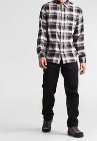 Koszula z flaneli w kratę w kolorach czarnym, białym i szarym, z dwoma kieszeniami na piersi i zapięciem na guziki, zestawiona z czarnymi spodniami i szarymi butami trekkingowymi.