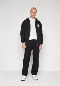 Svart zip-up hettegenser med hvit grafisk design på venstre bryst, kombinert med svarte bukser og hvite sneakers, som står mot en hvit bakgrunn.