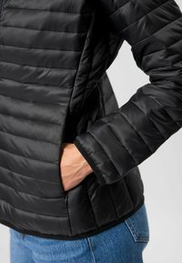 Zwarte gewatteerde jas met quilted textuur, staande kraag en zijzakken. Heeft een slim fit en rekbare mouwen. Draagt over blauwe jeans.