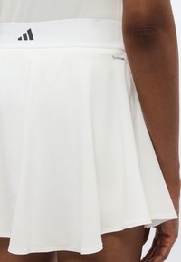 Λευκή φούστα τένις με ελαστική μέση, μαύρο λογότυπο Adidas, ελαφρύ ύφασμα και φαρδιά σχεδίαση για εύκολη κίνηση.