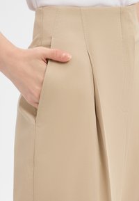 Beige tailored trousers with a high waist, featuring side pockets, pleats, and subtle stitching detail, made from a smooth fabric.